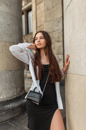 Fashion pretty young woman in stylish clothes with white shirt, black dress and handbag stands near a vintage building with columnsの写真素材