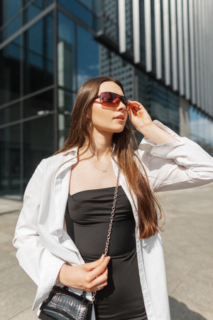 Street pretty beautiful youth stylish girl in fashion outfit with a black dress, a white shirt and a handbag walks on a sunny day in the city and puts on sunglassesの写真素材