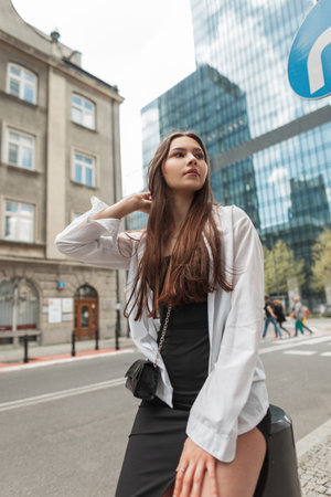 Stylish beautiful young lady in a fashion black dress with a white shirt with fashion bag sits in the cityの写真素材