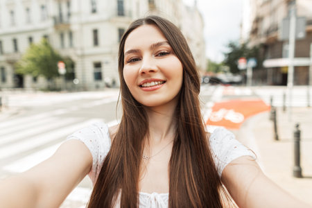 Happy beautiful young girl with a smile in a fashion white dress walks in the city on a sunny day and takes a selfie photo on a smartphoneの写真素材