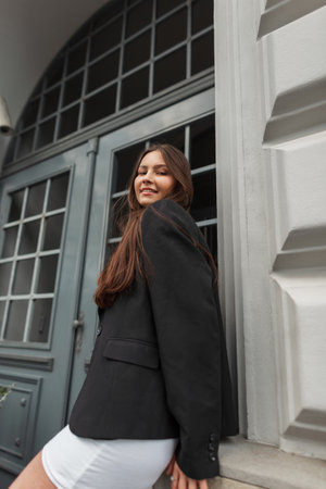 Urban fashion trendy happy pretty lady with stylish black leather and white dress stands near a vintage buildingの写真素材