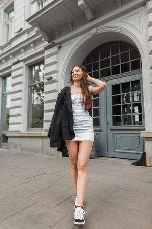 Happy fashionable young beauty woman with smile in casual street outfit with white dress, black blazer and shoes walks in the city near a vintage buildingの写真素材