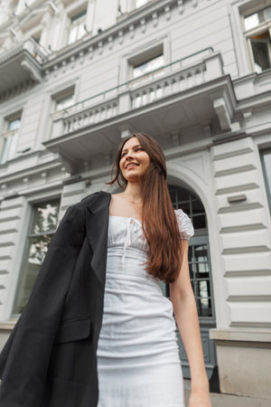 Happy beautiful fashionable girl model with smile in trendy fashion outfit with a white dress and a black blazer walks on the street near a vintage buildingの写真素材