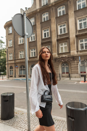 Fashion beautiful young woman model in stylish clothes in a black dress with a white shirt and a bag walks in the cityの写真素材
