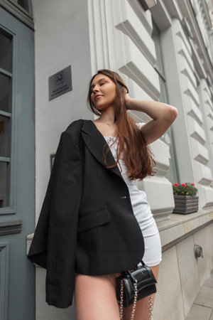 Fashionable young beautiful woman model in fashion clothes with white dress with black stylish blazer and handbag stands near a vintage white building outdoorsの写真素材