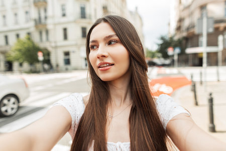 Beautiful fashion stylish girl in a fashionable white dress makes a selfie photo on a smartphone and walks in the cityの写真素材
