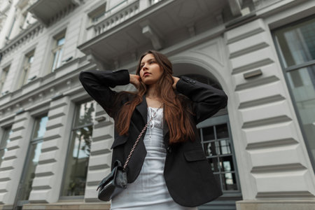 Beautiful young stylish girl model in a white dress with a black blazer with a handbag walks in the city near a vintage buildingの写真素材