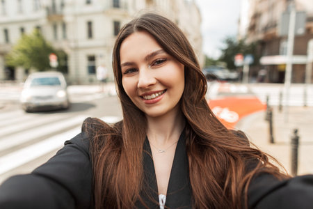 Happy beautiful young chic lady with a smile in fashion clothes with a black blazer walks in the city and takes a selfie on the cameraの写真素材