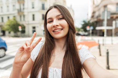 Happy beautiful young woman with a smile in a fashionable white dress walks in the city, takes a selfie photo on a smartphone and shows a peace signの写真素材