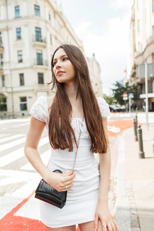 Pretty stylish fashion woman in fashionable white dress with black leather small bag walks in the cityの写真素材