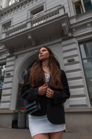 Beautiful serious young fashionable girl in stylish casual clothes with white dress and black blazer and handbag walks in the city near a white vintage buildingの写真素材