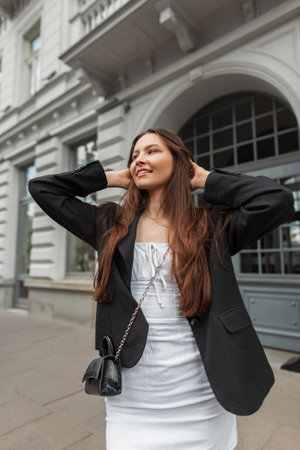 Fashionable young pretty happy woman with smile in stylish outfit with white dress and black blazer with handbag walks in the cityの写真素材