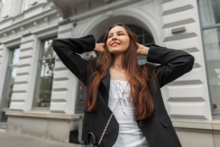 Happy young beautiful girl with a smile in stylish clothes with a white dress and a black blazer walks in the city near a vintage buildingの写真素材