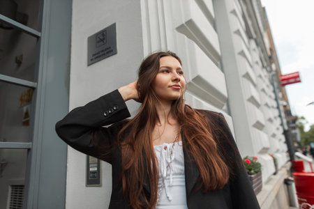 Happy fashion brunette woman model in stylish casual outfit street with white dress and black blazer walks in the city near a vintage buildingの写真素材