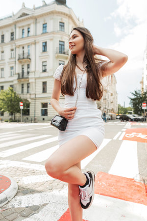 Beautiful cheerful young woman in fashion clothes in a white trendy dress with a stylish black bag and sneakers is walking and having fun in the cityの写真素材
