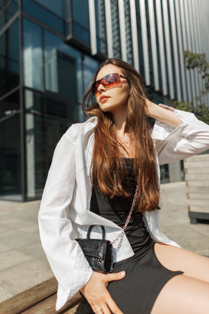 Fashionable beautiful chic girl with sunglasses in stylish urban outfit with a black dress and a white shirt with a handbag sits and poses in the city on a sunny dayの写真素材