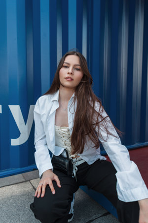 Fashionable beautiful youth woman model in trendy clothes with a white shirt with a stylish handbag sits and poses on the street near the blue metal backgroundの写真素材