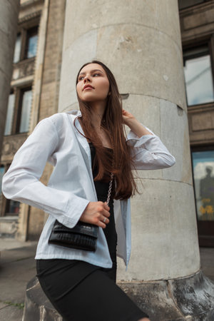 Stylish beautiful young woman model in casual clothes with a white shirt, black dress with a handbag walks in the city near a vintage building with columnsの写真素材