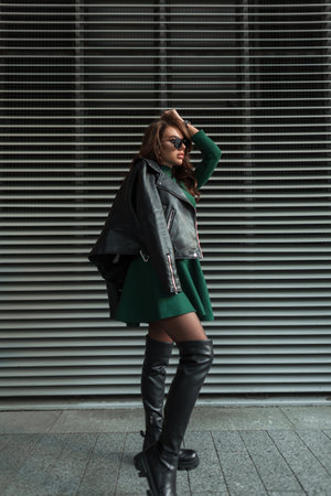 Glamor beautiful fashion girl with stylish sunglasses in fashionable rock clothes with a green dress with a leather black jacket in high shoes stands near a metal background on the streetの写真素材