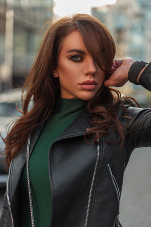 Pretty fashion lady with makeup in fashionable street clothes with black leather jacket and green sweater walks in the city and looks at the cameraの写真素材