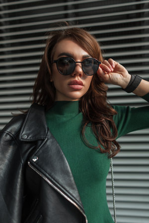 Pretty cool stylish beauty woman in trendy fashion clothes with black leather rock jacket puts on sunglasses and walks on the street near the metal lines backgroundの写真素材