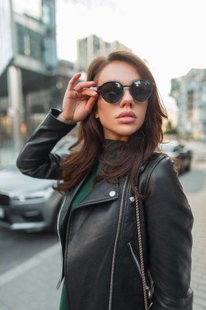 Beautiful fashion young stylish woman model in trendy clothes with a green dress and a fashionable leather black jacket walks in the city and puts on sunglassesの写真素材