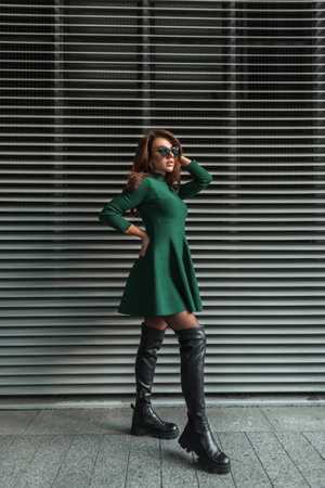 fashionable beautiful young girl with trendy sunglasses in a fashion green dress and stylish high leather shoes stands near the metal lines wall on the streetの写真素材