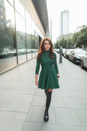 Beautiful fashion stylish girl in a trendy elegant green dress with a handbag and high leather boots walks in the cityの写真素材