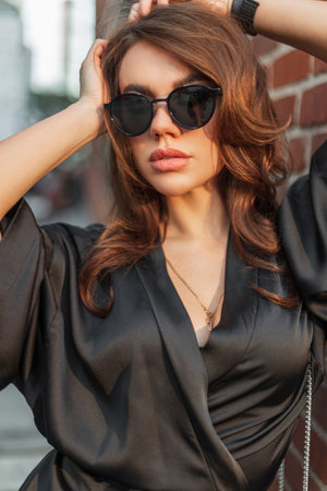 Fashionable beautiful stylish chic girl model with fashion trendy sunglasses in a stylish black elegant dress poses in the city near a brick wallの写真素材
