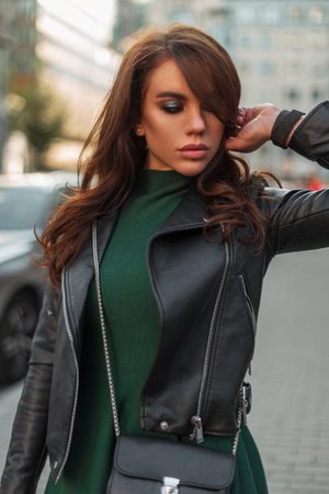 Stylish beautiful young woman model with makeup in fashion outfit with a leather jacket and a fashionable green dress with a trendy stylish handbag walks in the cityの写真素材