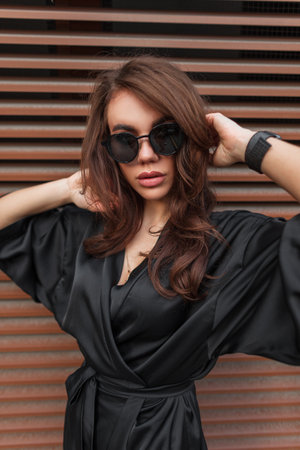 Fashion beautiful young chic woman model with a brunette hairstyle with stylish glasses in an elegant black dress on the street near a metal wallの写真素材
