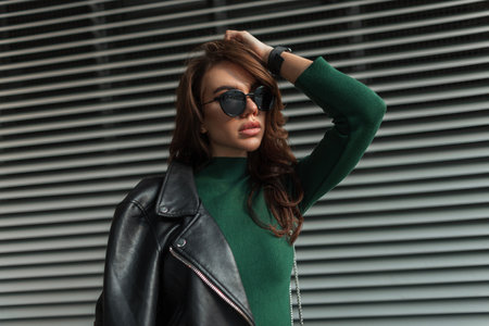 Stylish beautiful young lady with fashion sunglasses in trendy cool rock clothes with black leather jacket and green dress on the street near metal lines backgroundの写真素材