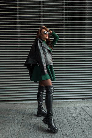 Cool beautiful stylish sexy woman with sunglasses in trendy fashion elegant rock outfit with green dress and black leather jacket with high shoes posing near metal wall outdoorsの写真素材