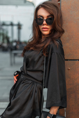 Fashionable young elegant pretty woman model with fashion sunglasses in elegant black suit with handbag stand near a wall in the cityの写真素材