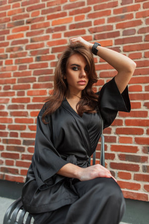 Fashionable beautiful young stylish chic woman with makeup in an elegant black suit sits near a brick wall. Pretty glamor ladyの写真素材