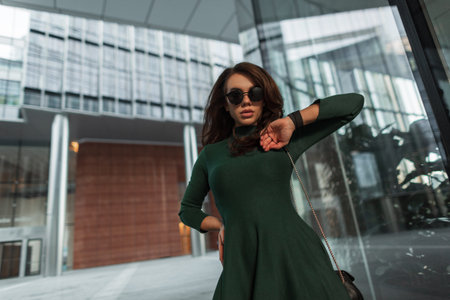 Beautiful fashionable cool pretty chic woman model with fashion black sunglasses in a stylish trendy green dress with a handbag walks in the city near the modern glass business buildingの写真素材