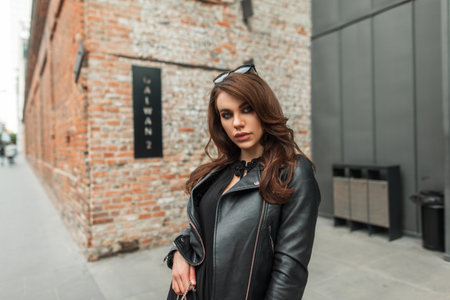 Beautiful young fashion woman model with sunglasses in trendy fashionable black clothes with a leather jacket and dress walks in the city near the buildingの写真素材