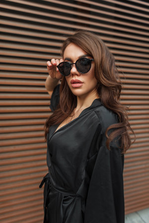 Fashionable beautiful elegant lady in a stylish black dress puts on sunglasses on the background of a metal rusty wall on the streetの写真素材