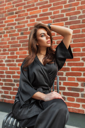 Beautiful fashionable stylish woman model with a hairstyle in a fashion elegant black suit sits near a red brick wall on the streetの写真素材