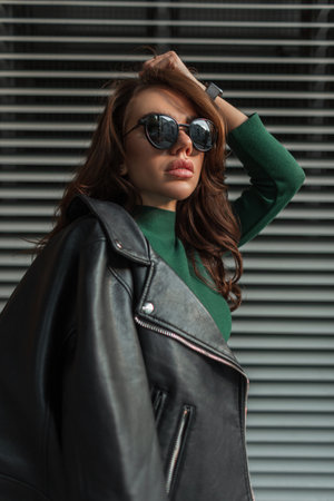 Fashionable young beauty woman with sunglasses in green dress with black leather jacket walks on the street near a metal lines wallの写真素材