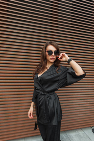 Glamorous beautiful fashion chic young woman model with brunette hair in a fashionable elegant black suit against the background of a metal rusty wall in the cityの写真素材
