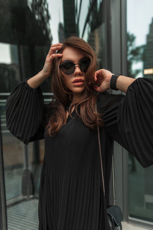 Urban female fashion portrait of stylish beautiful fashionable woman with cool vintage black sunglasses in trendy dress with small bag walks and poses on the street.の写真素材