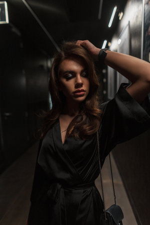 Fashionable beautiful chic woman in a stylish elegant black dress with bag in the dark indoorsの写真素材