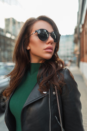 Urban beautiful young fashionable woman with cool vintage sunglasses in trendy rock street outfit with stylish black leather jacket and green dress walks in the cityの写真素材