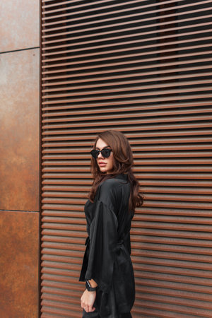 Fashion pretty beauty chic lady model with cool sunglasses in black fashionable dress walks near a rusty metal wall on the street. fashion style and outfitの写真素材