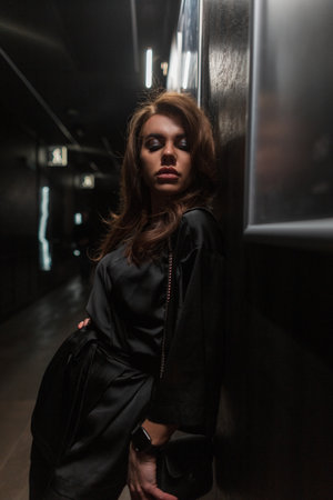 Beautiful fashionable young woman in an elegant fashion black dress stands near the wall in the dark indoorsの写真素材