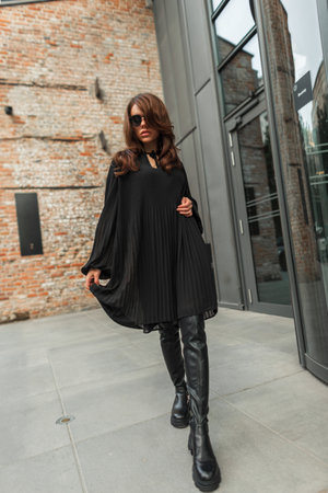 Glamorous beautiful fashionable woman model with stylish sunglasses in trendy fashion black outfit with a black fashion dress and high leather shoes walks in the city near a vintage buildingの写真素材