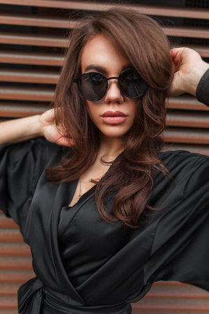 Outdoor female portrait of a beautiful stylish brunette woman with cool fashion sunglasses in a black dress near a metal wall in the cityの写真素材