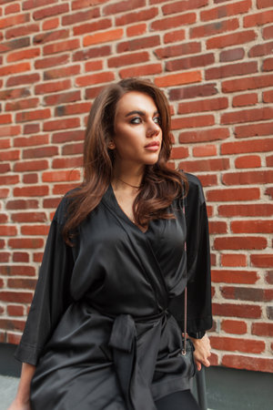 Fashionable beautiful young chic woman model in elegant black suit sitting near brick vintage wallの写真素材