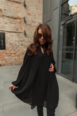 Beautiful stylish young woman model brunette with sunglasses in an elegant black fashion dress walks in the city near a vintage building. Pretty chic ladyの写真素材